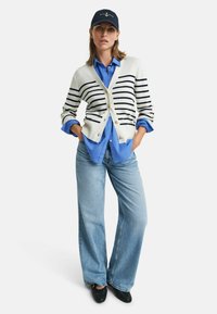 Cardigan rayé en crème et bleu marine, boutonné, superposé sur un chemisier bleu. Un pantalon large en denim bleu clair et des chaussures noires à enfiler complètent le look.