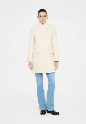 Manteau beige clair avec un col haut, au design structuré, poches avant et texture lisse. Assorti à un jean évasé bleu clair et des bottes noires.