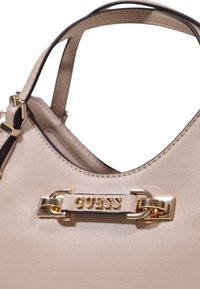 Bolso de cuero beige con herrajes dorados, que presenta un logo destacado de "Guess" y acentos negros en las correas y bordes. Textura suave.