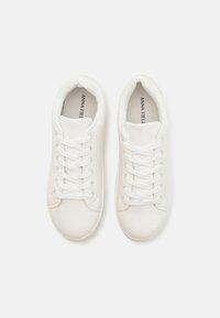 Des baskets blanches en matériau synthétique lisse présentent un bout rond, des lacets plats et des détails de couture subtils avec un design minimaliste.