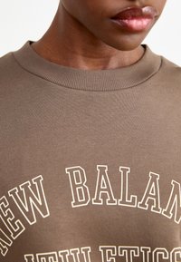 Brown cotton sweatshirt featuring a ribbed neckline and a large, bold cream-colored "NEW BALANCE" print.