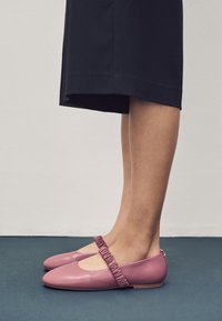 Roze leren ballerina's met een geplooid elastisch bandje over de wreef, een ronde neus en een platte zool, geplaatst op een donkerblauwe ondergrond.