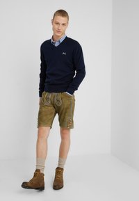Marineblauer Pullover über einem karierten Hemd, kombiniert mit khakifarbenen Shorts, die mit gestickten Mustern und Knöpfen verziert sind, abgerundet mit braunen Wanderstiefeln.
