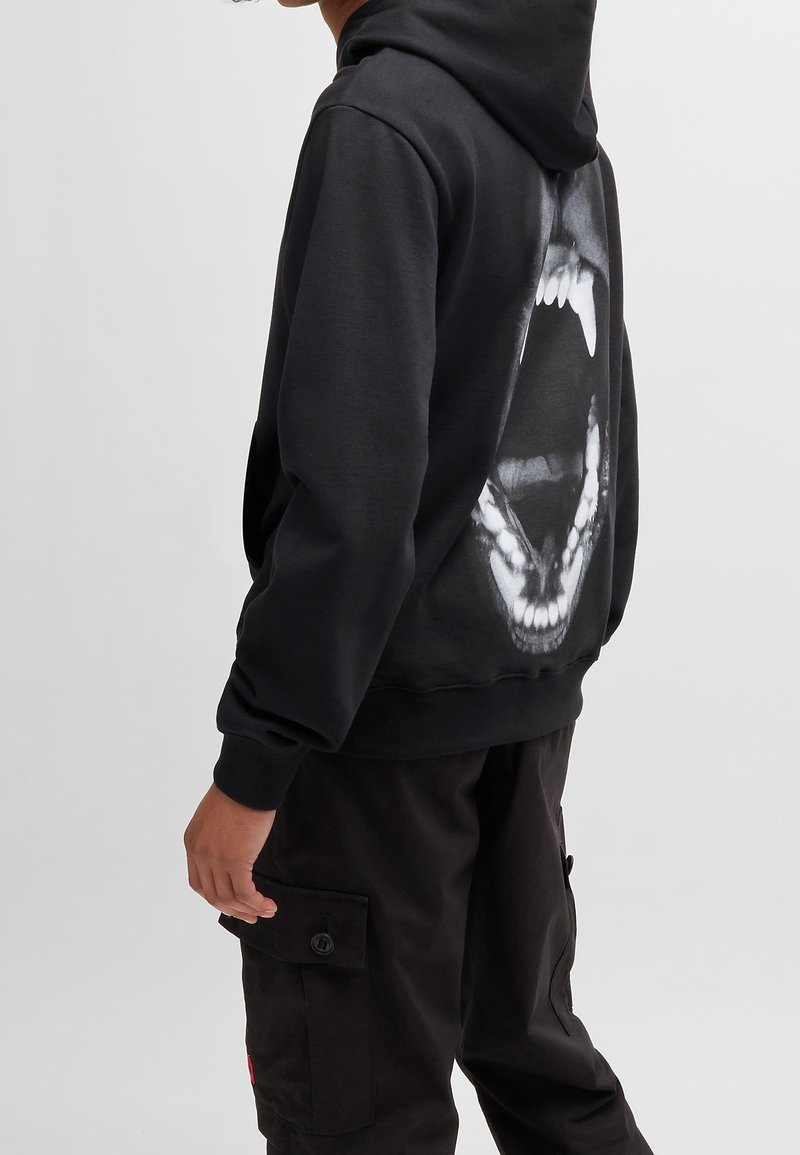 Personne portant un sweat à capuche noir avec un grand motif blanc d'une bouche d'animal ouverte au dos, associé à un pantalon cargo noir.