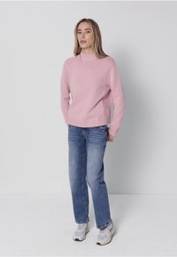 Hellrosa gestrickter Pullover mit hohem Kragen, kombiniert mit hellblauen, geraden Jeans und grauen Sneakern. Weiche Textur des Stoffes deutlich erkennbar.