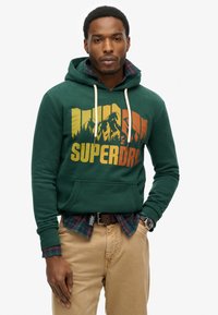 Zielona bluza z kapturem z graficznym nadrukiem w pomarańczowym i żółtym kolorze, z kieszenią kangurką i sznurkami. Nałożona na koszulę w kratę.