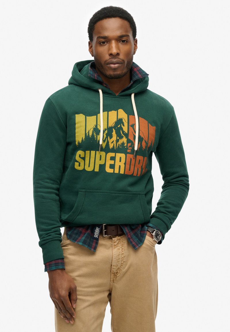 Zielona bluza z kapturem z graficznym nadrukiem w pomarańczowym i żółtym kolorze, z kieszenią kangurką i sznurkami. Nałożona na koszulę w kratę.