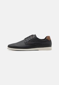Zapatos de cuero perforado negros con punta redondeada, acento en el talón marrón y suela blanca. Presenta cordones negros y un diseño minimalista.