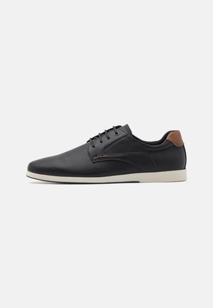 Black perforated leather shoes with a rounded toe, brown heel accent, and white sole. Features black laces and minimalistic design.
