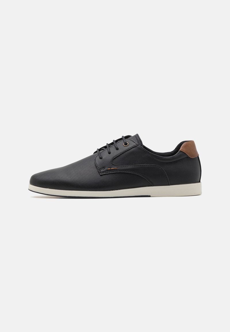 Zapatos de cuero perforado negros con punta redondeada, acento en el talón marrón y suela blanca. Presenta cordones negros y un diseño minimalista.