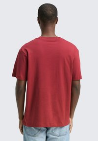 Młody mężczyzna widziany z tyłu, ubrany w gładką, czerwoną koszulę z krótkim rękawem i jasnoniebieskie dżinsy, na neutralnym tle.