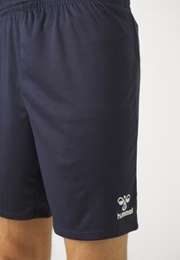 Mornarsko modri atletski kratke hlače z elastičnim pasom, ki jih nosi oseba, s belim logotipom Hummel blizu roba na desni nogi.