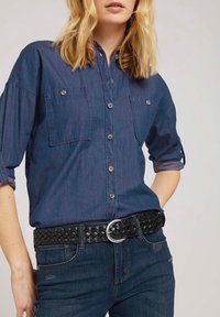 Camisa de mezclilla azul oscuro con dos bolsillos en el pecho y botones plateados, combinada con un cinturón trenzado negro que tiene una hebilla circular plateada.