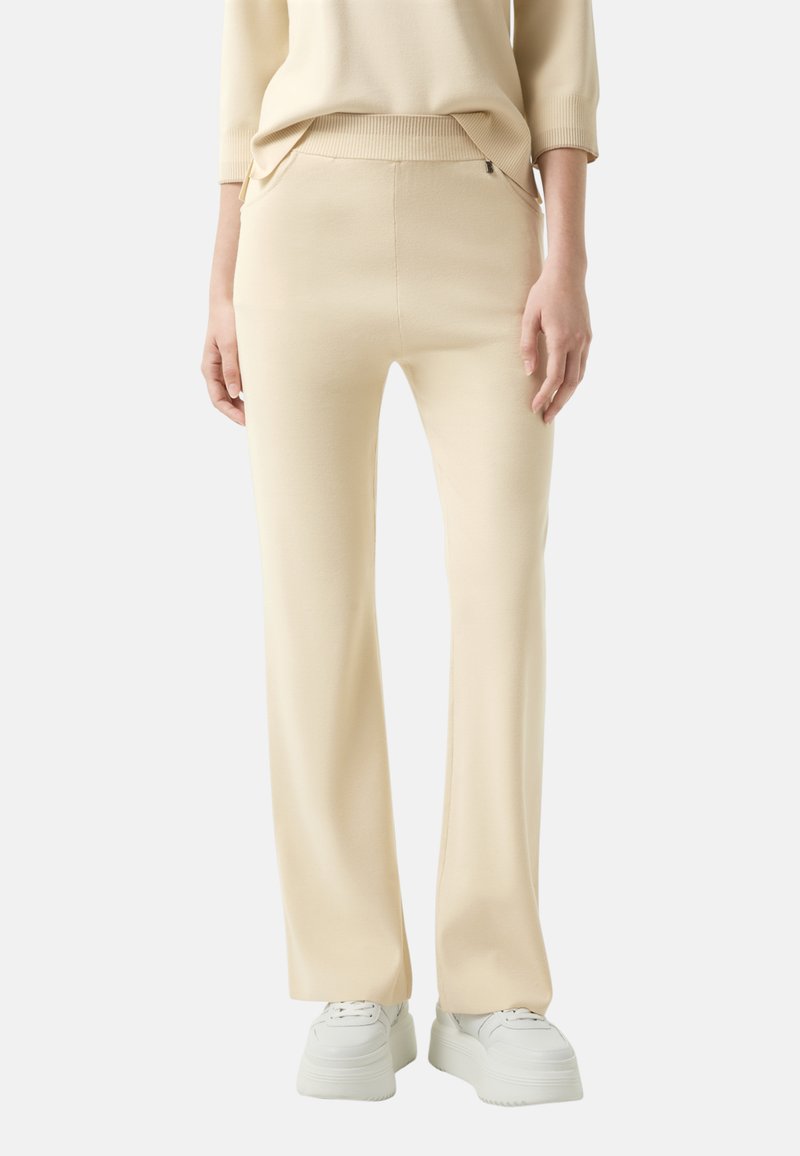 Cremfarbene Hose aus weichem, dehnbarem Material. Flared-Design mit glatter Textur und elastischem Bund. Verfügt über seitliche Taschen.