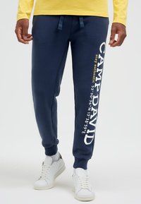 Mann trägt marineblaue Jogginghose mit der Aufschrift "CAMP DAVID" und Koordinaten auf dem rechten Bein, weiße Sneaker und ein gelbes Langarmshirt.