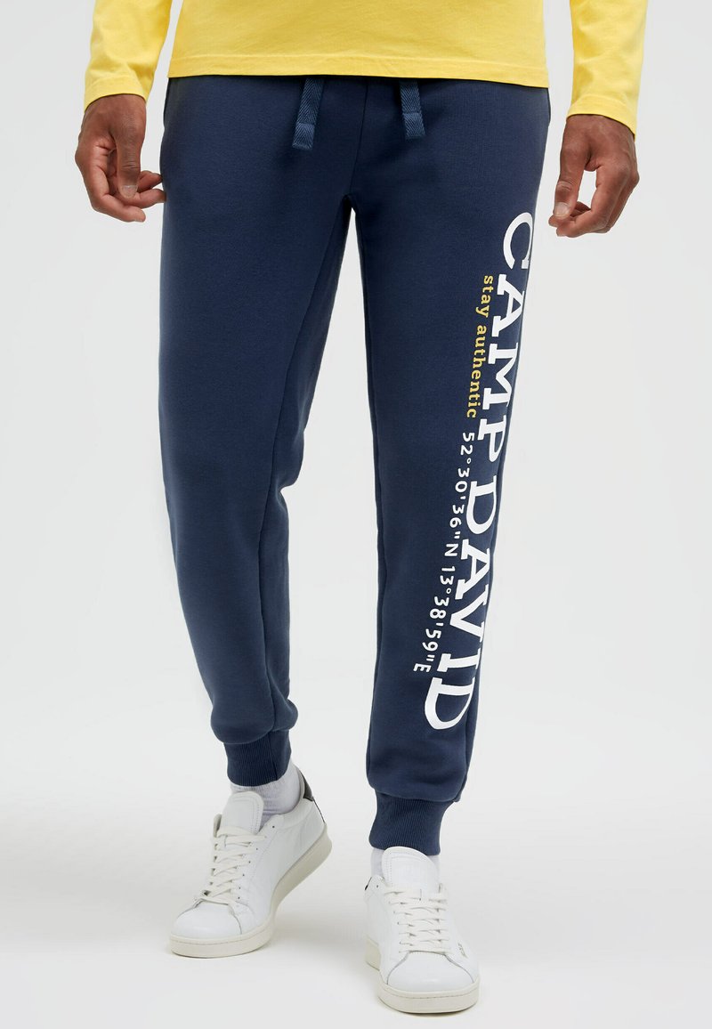 Mann trägt marineblaue Jogginghose mit der Aufschrift "CAMP DAVID" und Koordinaten auf dem rechten Bein, weiße Sneaker und ein gelbes Langarmshirt.