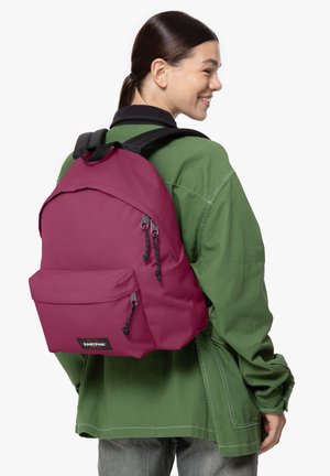 Mochila fucsia con una textura suave, que presenta correas ajustables negras, un bolsillo frontal y un parche de logo, llevada sobre una chaqueta verde.