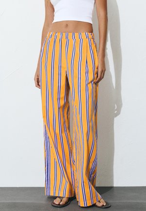 Kobieta ubrana w żółte spodnie z szerokimi nogawkami w pionowe niebiesko-białe paski, biały krótki top i czarne klapki, stojąca przy białej ścianie.