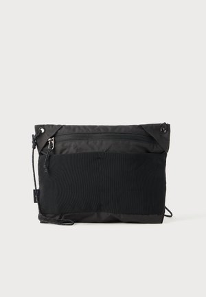 Borsa a tracolla nera con tasca superiore con zip, tasca frontale in rete e sottile tracolla regolabile su sfondo bianco semplice.