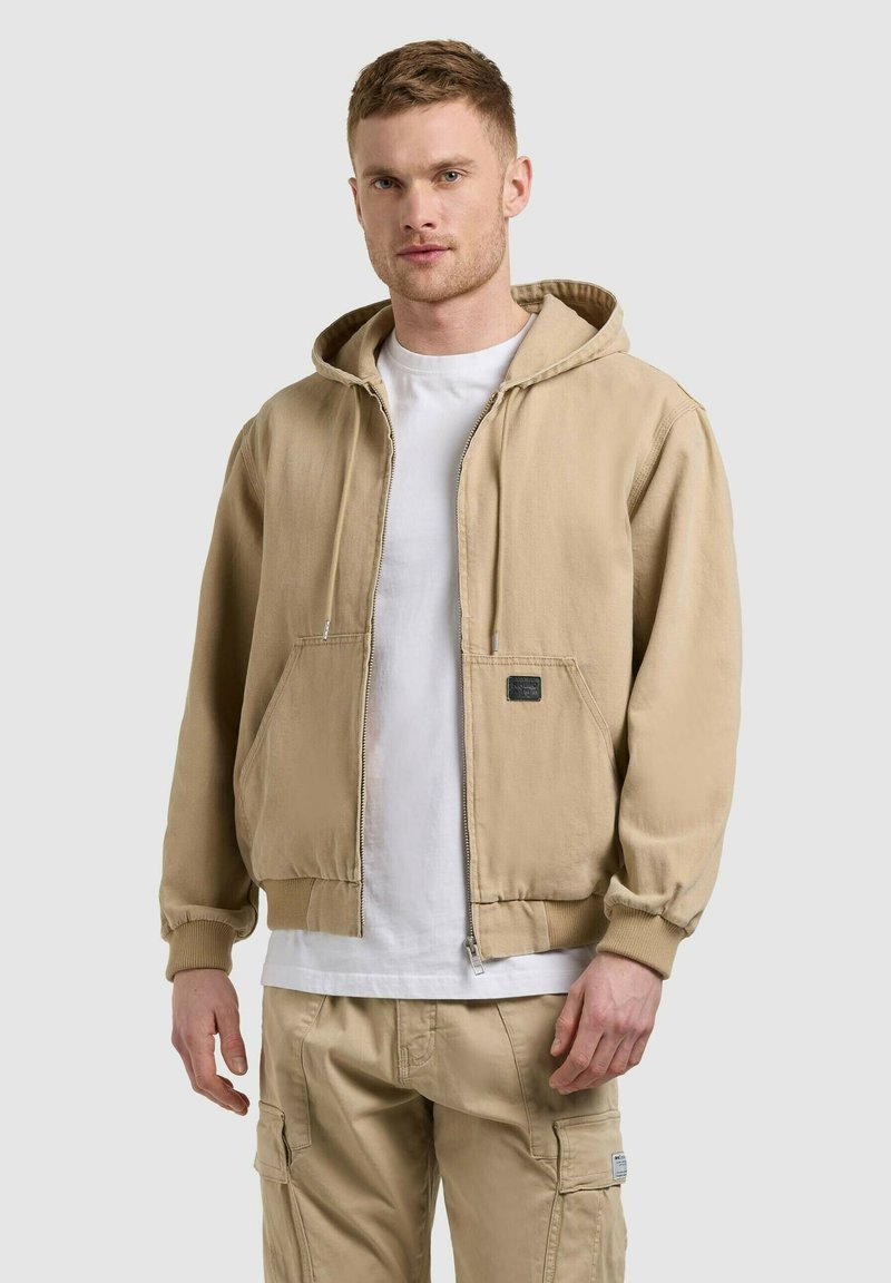 Homme portant une veste à capuche beige, un t-shirt blanc et un pantalon cargo beige, debout devant un fond uni gris clair.