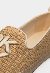 Μπεζ υφαντό παπούτσι χωρίς κορδόνια με παχιά σόλα και μεταλλικό λογότυπο "dk" στο μπροστινό πάνω μέρος.