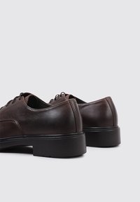 Chaussures en cuir marron avec une texture lisse, une forme arrondie et des semelles en caoutchouc noires. Présentent un design classique à lacets et des coutures minimales.