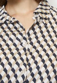 Camicia con bottoni caratterizzata da un motivo geometrico in beige, blu navy e bianco. Tessuto liscio con colletto classico e due bottoni visibili.