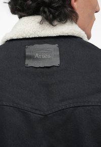 Persona con chaqueta negra y cuello de forro polar blanco, mostrando una patch de cuero negro etiquetada "Aries" en la parte superior de la espalda.