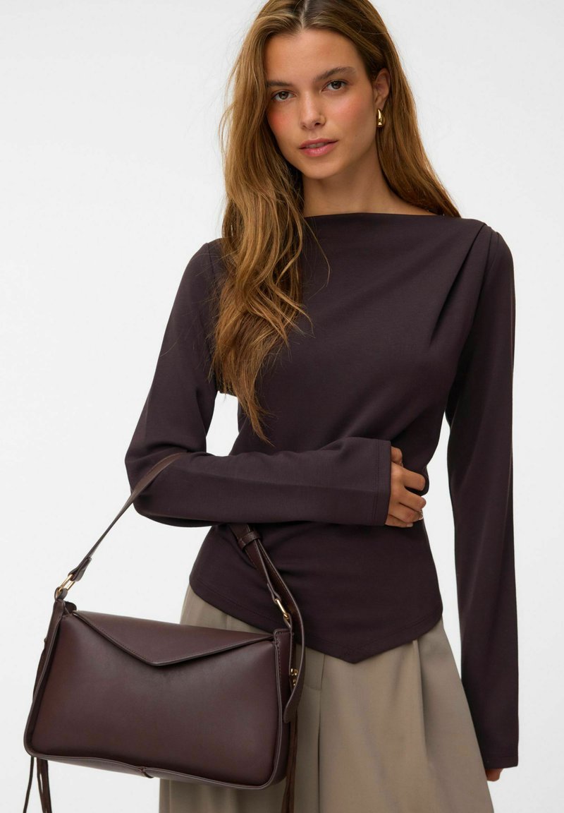 Femme aux cheveux longs portant un haut sombre à manches longues et une jupe beige, tenant un sac à main en cuir brun foncé sur son épaule.