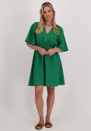 Groene jurk tot de generatie met een V-hals, korte uitlopende mouwen, een getailleerde taille en een gestructureerde stof. Draagt met bruine platte sandalen.