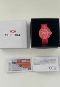 Rdeča silikonska ura s krožno obliko, srebrnimi označbami in logotipom "SUPERGA". Vključuje belo embalažno škatlo in kartico s podatki o produktu.