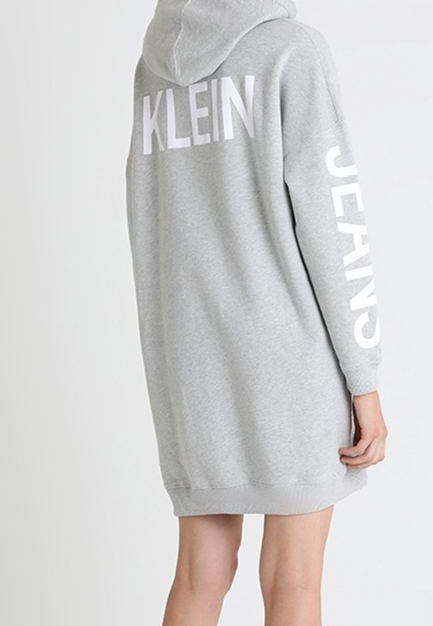 Personne portant une robe sweat à capuche gris clair avec « KLEIN » dans le dos et « JEANS » imprimé verticalement sur la manche.