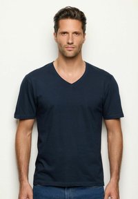 Muž s krátkými hnědými vlasy a světlým plnovousem, oblečený ve tmavě modrém tričku s výstřihem do V a modrých džínách, stojící proti jednolitému světlému pozadí.