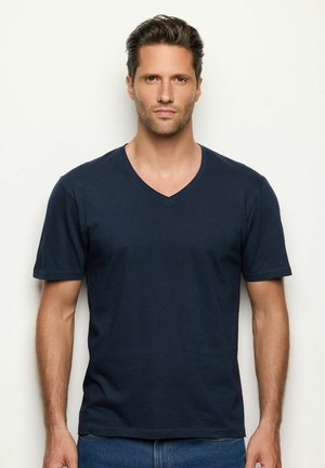 Hombre con cabello corto castaño y barba ligera, vistiendo una camiseta V azul oscuro y jeans azules, de pie contra un fondo claro y liso.