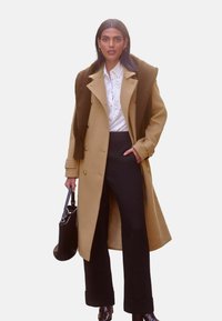 Beige Doppelreiher mit goldenen Knöpfen, texturiertem braunen Schal, weißer Bluse mit Spitzenverzierung und schwarzen weit geschnittenen Hosen. Schwarze Tasche.