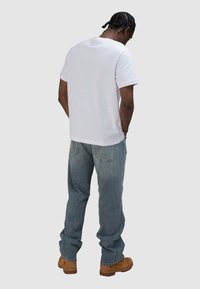 Witte katoenen T-shirt met korte mouwen, gecombineerd met lichtblauwe spijkerbroek met gedetailleerde stiksels op de achterzakken. Bruine schoenen zichtbaar.