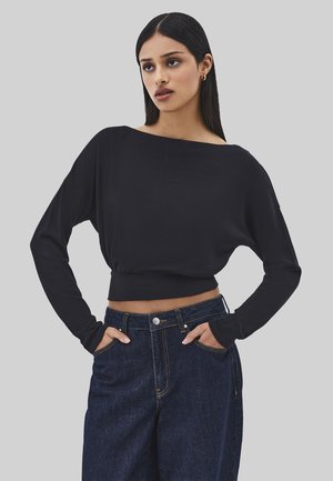 Jeune femme aux longs cheveux noirs portant un haut court noir à manches longues et un jean bleu foncé taille haute, les mains dans les poches.