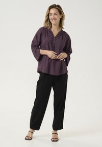 Lila Bluse mit V-Ausschnitt und Dreiviertelärmeln, versehen mit einer Rüschendetail und drei kleinen Knöpfen. Kombiniert mit schwarzen Hosen.