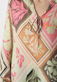 Bluse mit Blumenmuster und gebundenem Ausschnitt. Enthält geometrische Formen in Rosa-, Braun- und Grüntönen. Aus leichtem, strukturiertem Stoff gefertigt.