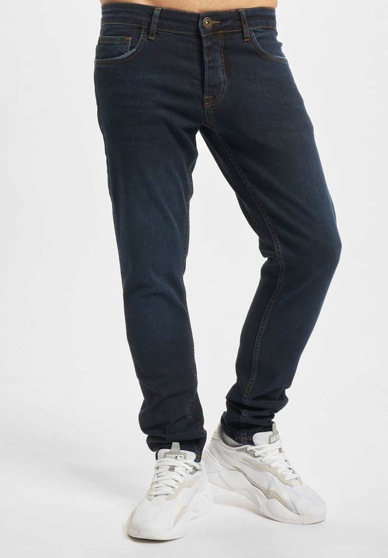 Homme portant un jean slim bleu foncé et des baskets blanches de sport, debout devant un fond uni.