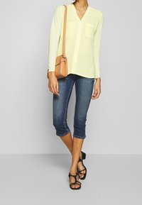 Gul långärmad blus med V-ringning och två framfickor, ihop med mörkblå caprijeans och svarta sandaler med remmar, bärande på en beige väska.