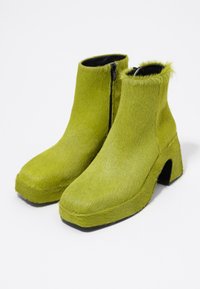 Botas de tobillo de peluche verde con puntera cuadrada, cierre de cremallera y un diseño de tacón recortado único. Textura suave y suela gruesa.