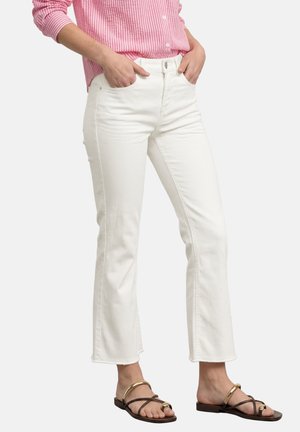 Personne portant un jean blanc évasé court, une chemise rayée rose à manches longues et des sandales marron avec des accents dorés, mains dans les poches.