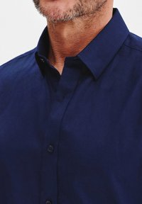 Granatowa koszula zapinana na guziki z kołnierzykiem, o gładkiej strukturze i subtelnym połysku. Posiada standardowe zapięcie na guziki i schludny fason.