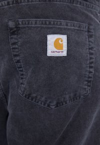 Tamno sivi kaput od kožne tkanine s bijelim Carhartt logotipom koji ima žute stilizirano "C" ušiveno na gornjem središtu.