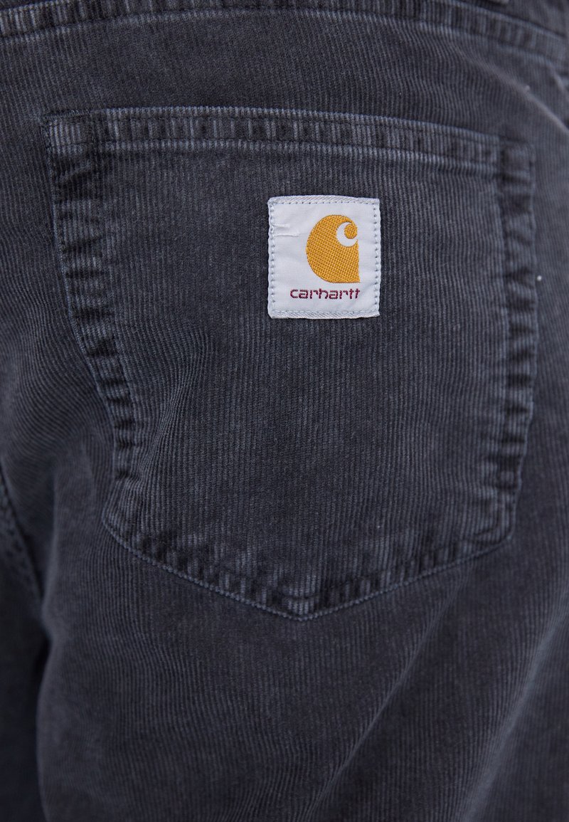 Mørkegrå kordfløjl bukselomme med et hvidt Carhartt logo patch, der har et gult stiliseret "C" syet på den øverste midte.