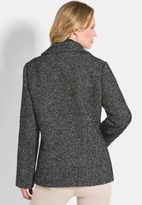 Vrouw met blond haar in een donkergrijze blazer van visgraatstructuur, staande met één hand op de heup, met haar rug naar de camera gekeerd.