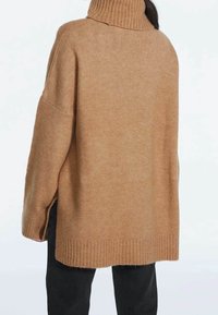 Pull à col roulé marron oversize avec poignets et ourlet côtelés, fabriqué dans une texture douce. Comprend des fentes latérales pour faciliter les mouvements.