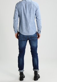 Helltürkises langärmeliges Hemd, lässige Passform, dunkelblaue Jeans, umgeschlagene Ärmel, schwarze Stiefeletten, einfarbiger Hintergrund.