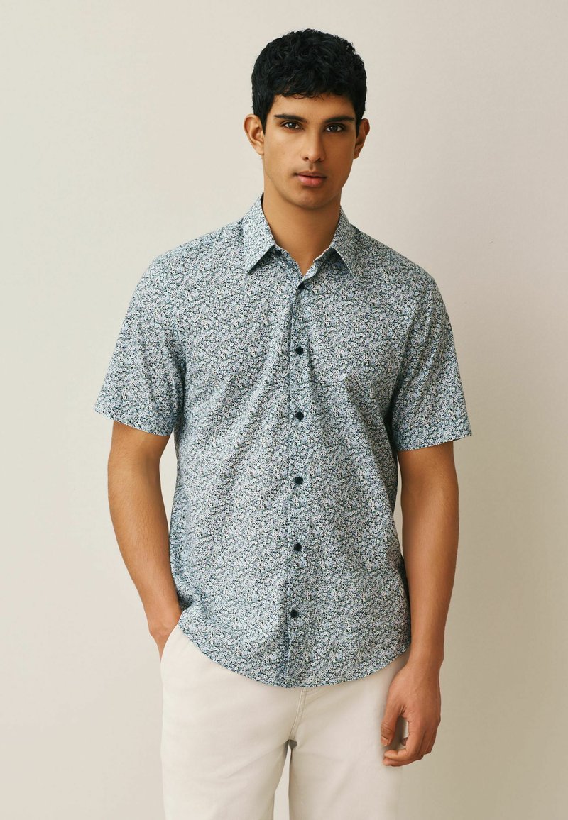 Jeune homme debout sur un fond clair uni, portant une chemise à manches courtes à boutons avec un petit motif floral et un pantalon clair.